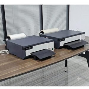 Two dual-head roll-fed desktop DTF transfer printers sitting side-by-side on a wooden workshop desk with pet film rolls mounted.